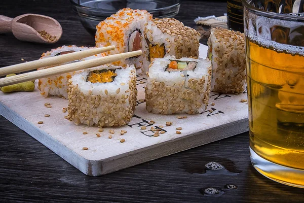 Sushi on a wooden plate with a glass of beer, soy sauce and natural wooden chopsticks on a light wooden table.