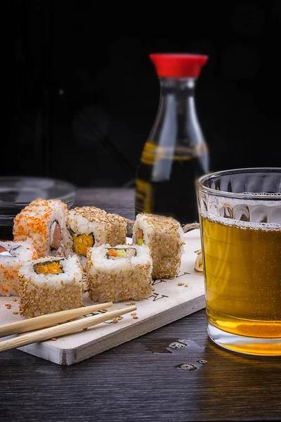 Sushi on a wooden plate with a glass of beer, soy sauce and natural wooden chopsticks on a light wooden table.