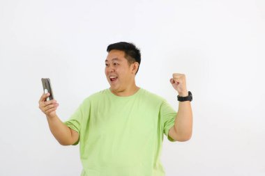 Genç Asyalı adam telefonu neşeli ve mutlu hareketlerle elinde tutuyor. Şimdi çok mutlu görünüyor. Başarı jestleri yapıyor.