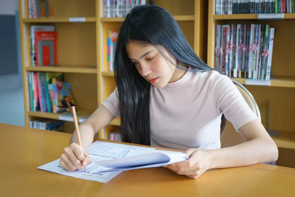 Young female university student concentrates doing language practice examination inside the library. Girl student writes the exercise of the examinations