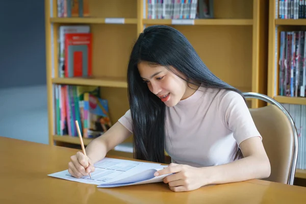 Young female university student concentrates doing language practice examination inside the library. Girl student writes the exercise of the examinations