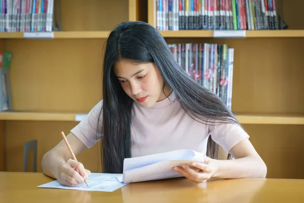 Young female university student concentrates doing language practice examination inside the library. Girl student writes the exercise of the examinations