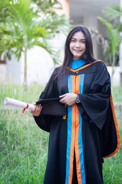 Mutlu ve heyecanlı Asyalı genç bayan üniversite mezunu portresi mezuniyet elbisesi giyer ve mezuniyet töreninde üniversite kampüsünde diplomayla kutlama yapar. Eğitim kavramı.
