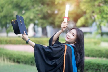 Mutlu ve heyecanlı Asyalı genç bayan üniversite mezunu portresi mezuniyet elbisesi giyer ve mezuniyet töreninde üniversite kampüsünde diplomayla kutlama yapar. Eğitim kavramı.