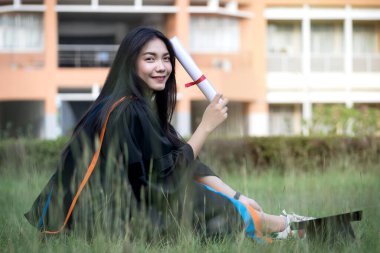 Mutlu ve heyecanlı Asyalı genç bayan üniversite mezunu portresi mezuniyet elbisesi giyer ve mezuniyet töreninde üniversite kampüsünde diplomayla kutlama yapar. Eğitim kavramı.