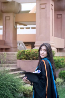 Mutlu ve heyecanlı Asyalı genç bayan üniversite mezunu portresi mezuniyet elbisesi giyer ve mezuniyet töreninde üniversite kampüsünde diplomayla kutlama yapar. Eğitim kavramı.