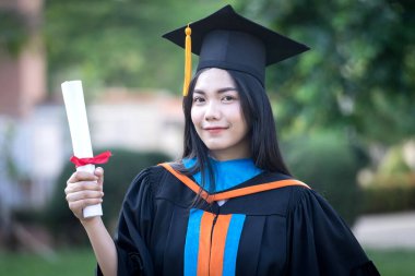 Mutlu ve heyecanlı Asyalı genç bayan üniversite mezunu portresi mezuniyet elbisesi giyer ve mezuniyet töreninde üniversite kampüsünde diplomayla kutlama yapar. Eğitim kavramı.