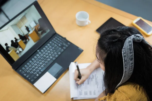 A teenager student studies online via laptop. University student girl watches online classes and writing a syllabus in notebook. Concept of distance study, online learning, webinars. Stock photo.