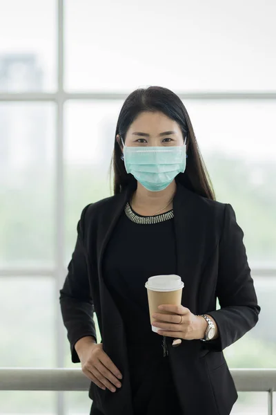 Portrait of a business woman with business suit wearing face mask holding morning coffee cup in company building ready for work. A middle age woman wears face mask to protect COVID-19 pandemic. Business stock photo
