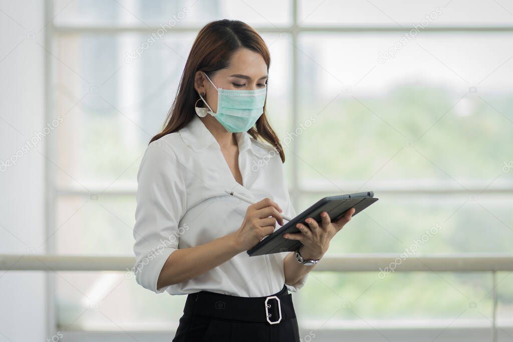 Portrait of a young adult Asian business woman in white shirt and black long pants wears surgical facemask looking on tablet in company building during VOCID-19 pandemic. Business stock photo.