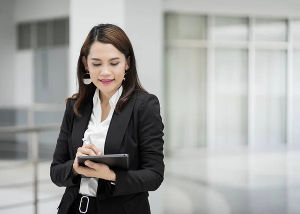 Portrait of a young cheerful businesswoman surfing social network on digital tablet in front of office during break. Asian business woman standing in office building. Stock photo