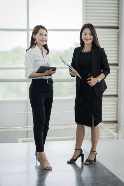 ビジネススーツを着た2人の女性オフィスワーカーが ビジネスビルでタブレットやファイルを使って仕事について話し合います ビジネスマンの女性が話し タブレットを指しています 業務用ストックフォト — ストック写真