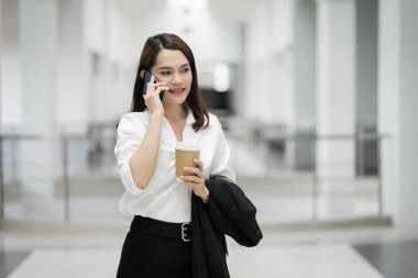 Genç Asyalı bir iş kadınının portresi cep telefonuyla konuşuyor ve iş binasında bir fincan kahve tutuyor. Güzel bir kızın resmi. Günlük süitte, telefon ve bir fincan kahveyle. Stok fotoğrafı