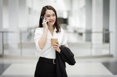 Genç Asyalı bir iş kadınının portresi cep telefonuyla konuşuyor ve iş binasında bir fincan kahve tutuyor. Güzel bir kızın resmi. Günlük süitte, telefon ve bir fincan kahveyle. Stok fotoğrafı