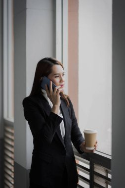 Genç Asyalı bir iş kadınının portresi cep telefonuyla konuşuyor ve iş binasında bir fincan kahve tutuyor. Güzel bir kızın resmi. Günlük süitte, telefon ve bir fincan kahveyle. Stok fotoğrafı