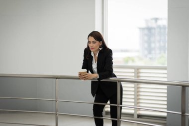 Mola sırasında iş yerinde cep telefonu kullanırken elinde bir fincan kahve tutan takım elbiseli kendine güvenen bir iş kadınının portresi. İş yeri fotoğrafı.