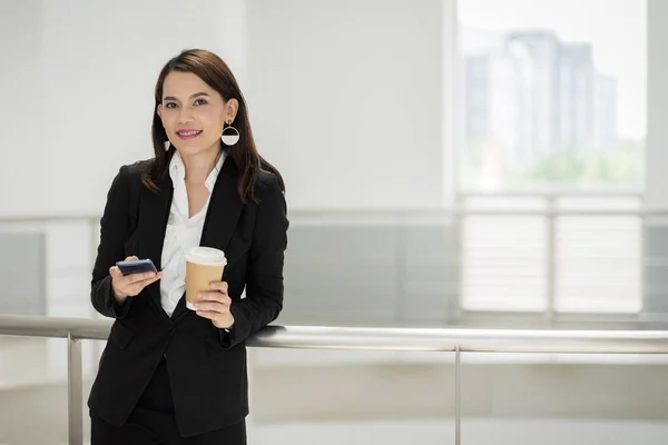 ビジネスビルで休憩時間に携帯電話を使用しながらコーヒーカップを保持しているビジネススーツに自信のあるビジネスマンの肖像画 業務用ストックフォト — ストック写真