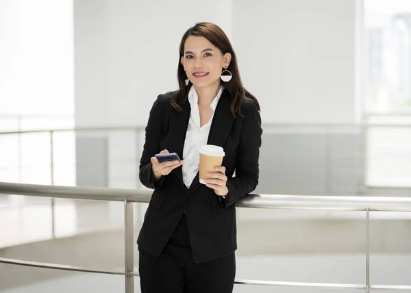 ビジネスビルで休憩時間に携帯電話を使用しながらコーヒーカップを保持しているビジネススーツに自信のあるビジネスマンの肖像画 業務用ストックフォト — ストック写真