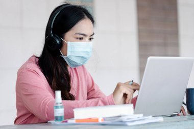 Coronavirus ya da COVID-19 salgını sırasında evde dizüstü bilgisayarda çalışan yüz maskeli genç Asyalı iş kadını ev konseptinden çalışıyor. Stok fotoğrafı