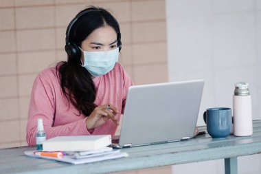 Coronavirus ya da COVID-19 salgını sırasında evde dizüstü bilgisayarda çalışan maskeli genç bir kadın evde online çalışan bir ault öğrencisidir. Stok fotoğrafı