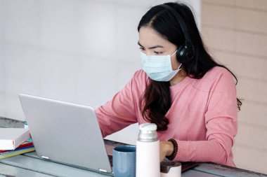 Coronavirus ya da COVID-19 salgını sırasında evde dizüstü bilgisayarda çalışan maskeli genç bir kadın evde online çalışan bir ault öğrencisidir. Stok fotoğrafı