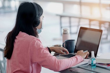 Coronavirus ya da COVID-19 salgını sırasında evde dizüstü bilgisayarda çalışan maskeli genç bir kadın evde online çalışan bir ault öğrencisidir. Stok fotoğrafı