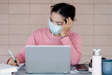 Coronavirus ya da COVID-19 salgını sırasında evde dizüstü bilgisayarda çalışan maskeli genç bir kadın evde online çalışan bir ault öğrencisidir. Stok fotoğrafı