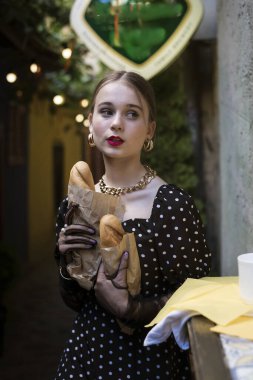 Çekici genç bir kadının portresi. Eski bir kasabada, dokulu bir duvarda baget yiyor. Beyaz benekli klasik siyah elbise, ellerinde siyah eldiven, düz saçlı..