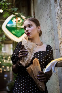 Sevimli genç bir kadın, eski bir şehrin desenli duvarının arka planında baget yiyor. Beyaz puantiyeli siyah bir elbise, ellerinde siyah eldivenler ve pürüzsüz bir saç stili..