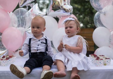 İki çocuk, erkek ve kız kardeş, ikizler ilk doğum günlerini kutluyorlar. Balonların arka planına karşı giyinmiş, yan yana oturuyorlar. Çocuk Partisi Açık havada.