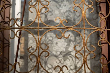 Zengin dekore edilmiş demir ızgara ve eskimiş ahşap çerçeveli, soluk beyaz perdeli, huzur ve sükunet ortamı yaratan tarihi bir pencere.