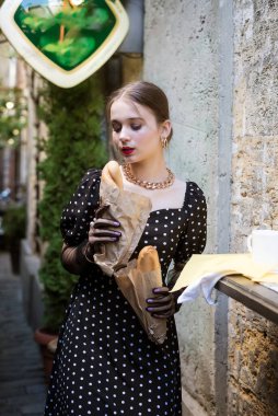 Sevimli genç bir kadın, eski bir şehrin desenli duvarının arka planında baget yiyor. Beyaz benekli siyah bir elbise, ellerinde siyah eldivenler ve pürüzsüz bir saç stili..