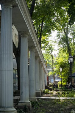 Parktaki yeşilliklerin etrafında bir sıra beyaz sütun. Yapı zarif sütunlarla süslenmiş ve sessiz bir yolun yanında huzurlu bir dinlenme sunuyor..