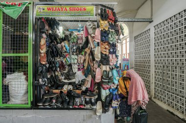 Sandaletler, spor ayakkabıları ve resmi ayakkabılar da dahil olmak üzere çeşitli ayakkabıları sergilenen küçük bir ayakkabı dükkanı. Pembe başörtülü bir kişi seçime göz atıyor.