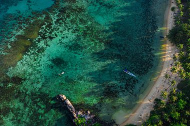 Berrak turkuaz suları, mercan resifleri ve palmiye ağaçları ve kayalık oluşumlarla çevrili tekneleri olan tropik bir sahil manzarası.