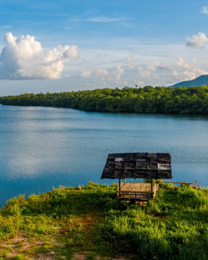 Bereketli yeşilliklerle çevrili ahşap bir çardağı olan sakin bir göl manzarası ve kabarık bulutlu mavi bir gökyüzünün altında sakin bir su.