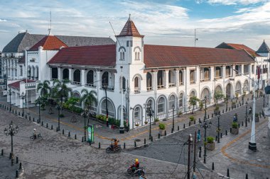 Kemerli pencereleri ve kırmızı çatısı olan tarihi sömürge binası, mavi gökyüzünün altında, kaldırım taşı sokak ve motosikletlerle çevrili..
