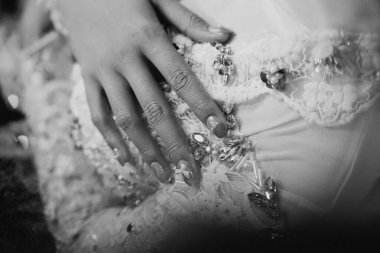 Dantel ve süslerle bezenmiş bir gelinliğin üzerinde dinlenen bir elin yakın çekimi. Karmaşık detaylar ve zarif tasarım sergileniyor.