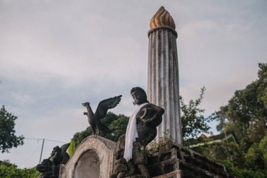 Bir kuştan oluşan heykel, arka planda yeşil bir alevle çevrili uzun bir sütunu olan bir kuşu tutuyor.