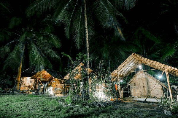 Спокойная ночная сцена с бамбуковыми хижинами в окружении пальм, освещенная мягкими огнями, демонстрирующая тропическую атмосферу