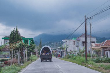 Bulutlu bir gökyüzünün altında evlerle ve yeşilliklerle çevrili bir kamyon taşıyan kırsal bir yol.