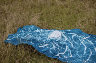 Mavi bir kumaş çimlerin üzerine serilmiş, beyaz soyut desenler ve metinler içeriyor, doğa ve sanat duygusunu çağrıştırıyor.