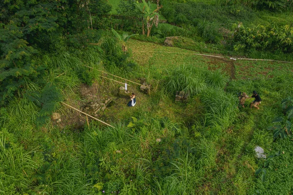 Yüksek otlu yemyeşil bir arazinin havadan görünüşü, su kaynağının yanında diz çökmüş bir kişi, ve arka planda ağaçlar ve tarım arazileriyle çevrili başka bir kişi.