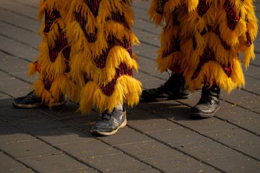 İki çift renkli, tüylü sarı kostümlü, kırmızı aksanlı, biri spor ayakkabılı, diğeri siyah çizmeli, kaldırımda duran.