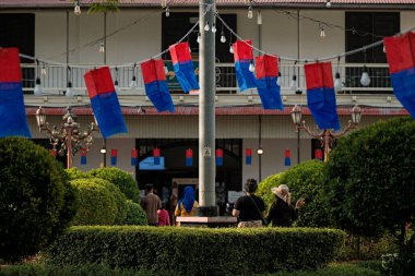 Kırmızı ve mavi bayraklarla süslenmiş bir halk meydanı, etrafta yürüyen insanlar, ön planda yeşillik, ortada bir bayrak direği, ve arka planda bir bina