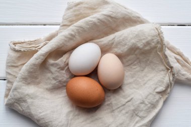 Beyaz, kahverengi ve bej olmak üzere üç farklı tavuk yumurtasının yukarıdan görünüşü beyaz ahşap bir yüzeye yerleştirilmiş keten peçetenin üzerinde duruyor. Kırsal çiftlik taze gıda konsepti