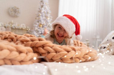 Tasarımcı battaniye merinos bej üzerinden! Noel Baba şapkası gülen çocuk. içinde belgili tanımlık geçmiş kız arkasında cam kenarında bir Noel ağacı var. mutlu çocuk Noel başlangıcında sevindirici yatakta yalan