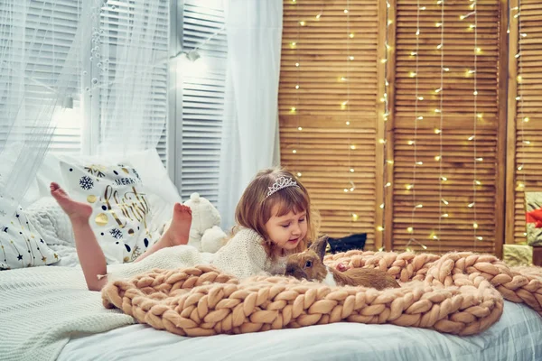 little girl in a knitted dress in bright color and the rim lies on the bed dangling bare feet on a soft designer plaid large knit of Merino, looking at the rabbit with a protruding ear. tint