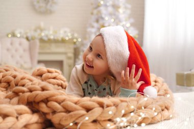 Tasarımcı battaniye merinos bej üzerinden! Noel Baba şapkası gülen çocuk. içinde belgili tanımlık geçmiş kız arkasında cam kenarında bir Noel ağacı var. mutlu çocuk Noel başlangıcında sevindirici yatakta yalan