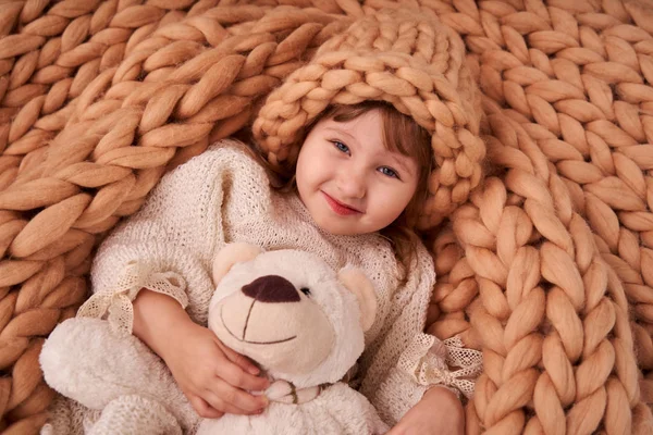 child in a hat under a blanket of natural sheep wool. Merino plaid knit covers little girl. designer hats natural wool yarn. crocheted dress. happy baby in a crib hugging a Teddy bear